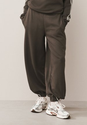 Persona que lleva puestos pantalones jogger sueltos de color marrón oscuro con puños elásticos y zapatillas de correr Nike blancas sobre un fondo liso.