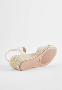 Sandal med kilehæl med en vævet jute sål, overdel af cremefarvet stof og justerbare stropper. Glat beige ydersål med struktureret grebmønster.