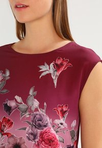Blouse florale bordeaux avec manches courtes, col rond et motifs floraux complexes en nuances de rose et de gris. Texture de tissu lisse.