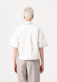 Camicia bianca a maniche corte con cuciture nere a contrasto lungo il colletto e i bordi delle maniche, dotata di una piega posteriore per aggiungere volume.