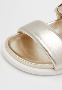 Närbild av den framre delen av en gyllene metallisk sandal med öppen tå, bred rem och vit sula på en vit bakgrund.