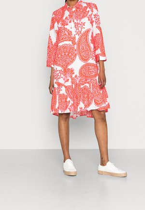 Kobieta w białej sukience do kolan z odważnym wzorem pomarańczowego paisley i białych trampkach z podeszwami z juty, stojąca na szarej podłodze.