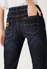 Vaqueros de mezclilla azul oscuro con un diseño ajustado. Presentan costuras amarillas y dos bolsillos traseros con una etiqueta de logo. Textura suave y perneras entalladas.