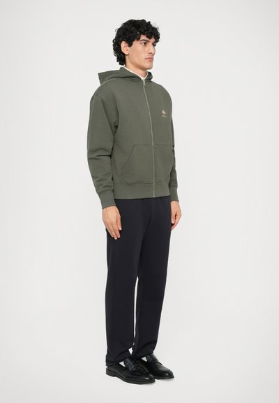 Hombre de pie, vestido con sudadera con cremallera color verde olivo y bolsillos frontales, pantalones negros y mocasines negros, contra un fondo blanco liso.