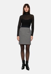 Black sheer turtleneck with polka dots paired with a fitted, patterned skirt in gray and black. Model wears black heeled ankle boots.