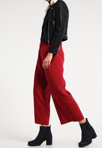 Chaqueta de motorista de cuero negro con cuello de piel sintética, pantalones rojos de pierna ancha y botines azul marino con tacón ancho y cremallera lateral.