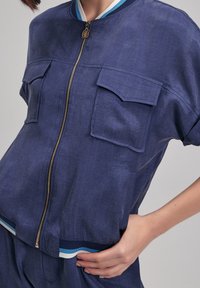 Granatowa kurtka typu cropped z krótkimi rękawami, dwoma przednimi kieszeniami, złotym zamkiem błyskawicznym oraz prążkowanym dołem z białymi i niebieskimi paskami.