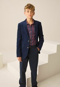 Tumedast värvitud korduroobi bleiser punase ja sinise ruudulise särgi peal, koos tumedate pükstega. Sile tekstuur ja lihtsad nupud.