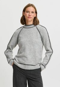 Pulover gri cu un guler înalt, tiv elastic și accente gri închis pe cusături. Textură moale, croială ușor lejeră, stilizat cu pantaloni închiși la culoare.