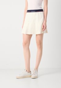 Lacoste plisserad kjol i off-white, med midjeband i marinblått och röda detaljer. Tillverkad av lätt material, designad för komfort och rörelse.