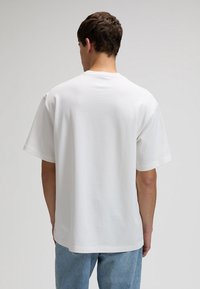 Junge Person, von hinten gesehen, trägt ein einfaches weißes kurzärmeliges T-Shirt und hellblaue Jeans vor neutralem Hintergrund.