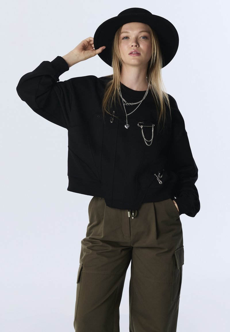Zwarte cropped sweatshirt met ritsdetails en zilverkleurige hardware, gecombineerd met olijfgroene cargobroek met zakken en een trekkoord in de taille.