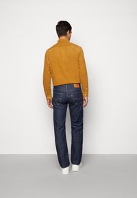 Man som bär en senapsfärgad manchesterjacka, mörkblå denimjeans och vita sneakers står vänd mot en enkel vit vägg.