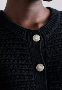 Mustaknitty cardigan, jossa on teksturoitu kuvio, pyöreä pääntie ja hopeasävyiset napit, joissa on kukkaembleemi kiinnityksessä.
