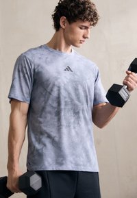 Pilkas tie-dye sportinis marškinėliai su trumpomis rankovėmis ir apvaliu kaklu, derinami su juodomis šortais. Juodi hanteliai laikomi kiekvienoje rankoje.