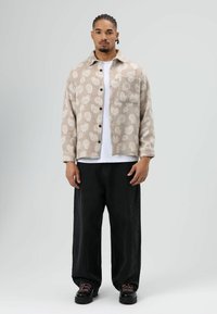 Beige overhemdjack met patroon, knopen en een borstzak, gedragen over een wit T-shirt. Zwarte broek met wijde pijpen en stevige zwarte schoenen.