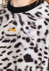 Top de estampado de leopardo con cuello negro y pequeño logo amarillo "RAW." cerca del hombro izquierdo, llevado por una persona.