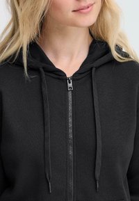 Svart zip-up hettegenser laget av mykt stoff, med snor i hetten, metallglidelås og ribbede detaljer. Nærbilde av halslinningen.