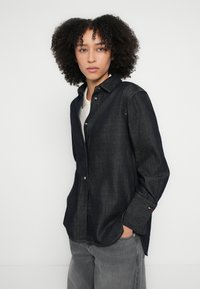 Σκούρο πουκάμισο από τζιν με κουμπιά, με μυτερό γιακά, μακριά μανίκια και διακριτική αντίθεση ραφών. Περιλαμβάνει λογότυπο στην μανσέτα.