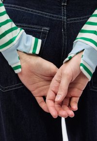 Μπλούζα με μακριά μανίκια με ρίγες σε πράσινο και λευκό, μπλε μανσέτες, χέρια σταυρωμένα μπροστά, σκουρόχρωμο φόντο από τζιν υλικό.