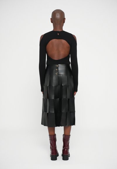 Persona vestida con una blusa negra de manga larga con un gran escote en la espalda, una falda midi negra texturizada con botones y botas de tobillo burdeos.
