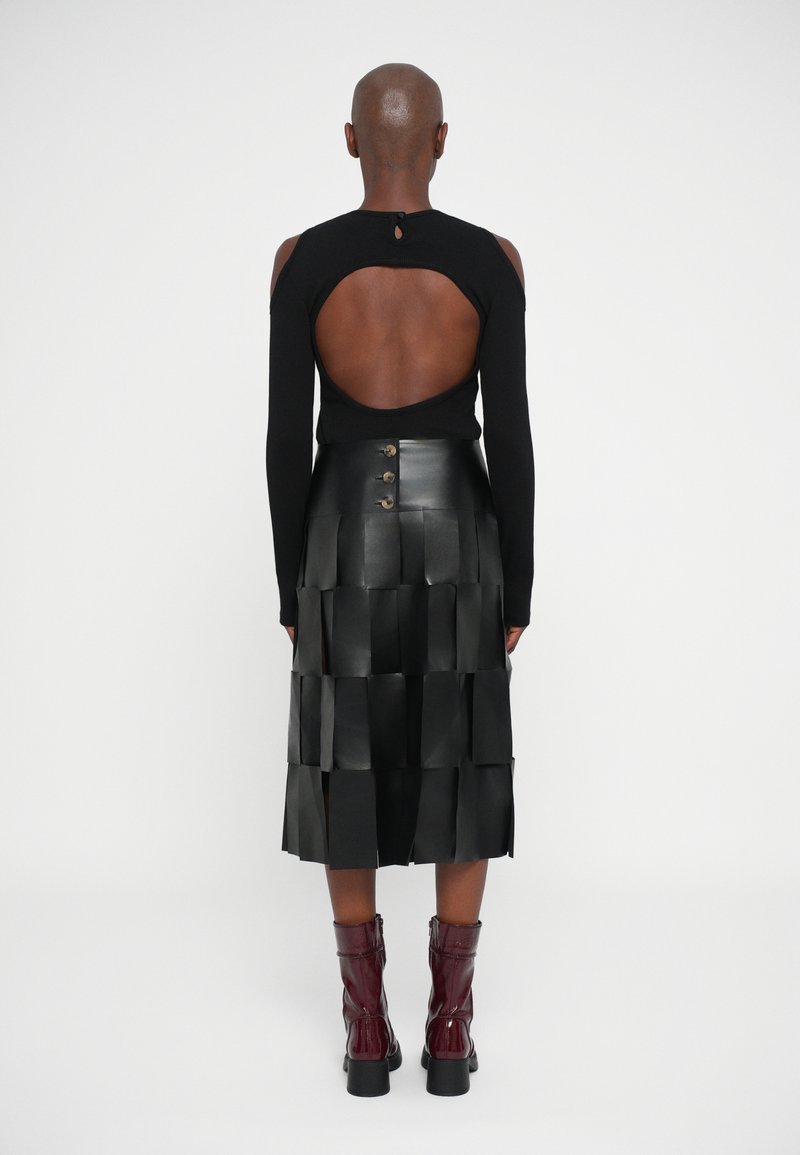 Persoon in een zwart lang mouwtop met een grote uitsparing op de rug, een zwarte gestructureerde midi-jurk met knopen en bordeauxrode enkellaarsjes.