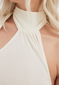 Primer plano de una blusa de tirantes color beige claro con detalles de tela fruncida, llevada por una persona con cabello largo y rubio.