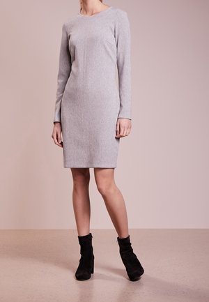 Femme portant une robe grise ajustée à manches longues et longueur genou, associée à des bottines noires, debout sur un sol lisse devant un fond uni.