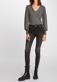 Kvinna som bär en kolgrå V-ringad långärmad topp med knäppning i ärmsluten, svarta högmidjade skinny jeans och svarta ankelskor.