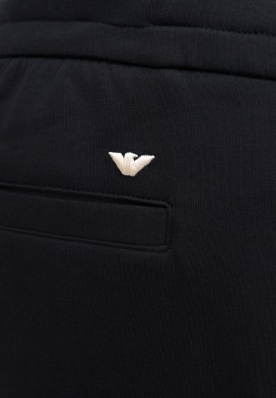 Κοντινή λήψη σε μαύρο παντελόνι από ύφασμα με οριζόντια πίσω τσέπη και ένα μικρό λευκό κεντητό λογότυπο αετού πάνω από την τσέπη.