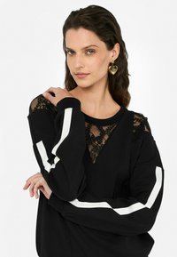 Sweatshirt noir avec des détails en dentelle sur les épaules et des accents de rayures blanches sur les manches. Tissu doux avec une coupe décontractée.