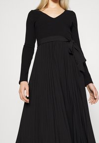 Sort ribbede strikkjole med lange ærmer og V-udskæring. Udfoldet, plisseret nederdel med et stofbælte bundet om taljen. Glat tekstur overalt.