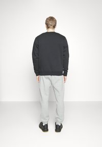 Hombre de pie frente a una pared blanca, vestido con sudadera negra, pantalones de chándal gris claro, calcetines blancos y zapatos negros.