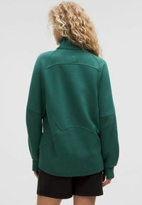 Pullover in pile verde con colletto alto, caratterizzato da una trama a costine, dettagli di cucitura e un logo sul retro. Abbinato a pantaloni corti neri.
