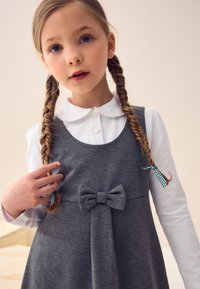 Ragazzina con capelli intrecciati legati da nastri a quadretti, che indossa una camicia bianca con colletto sotto un vestito grigio con un fiocco frontale.