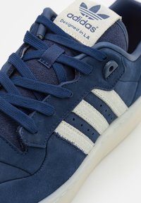 Sötétkék sneakers, velúr és hálós anyagból készült, három bézs csíkkal, párnázott nyelvvel és bordázott gumitalppal.