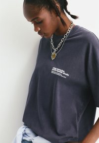 Mørkegrå bomulds t-shirt med korte ærmer og grafisk tekst. Båret med lagdelte sølv kædetrøjer og en lyseblå stribet accessory.