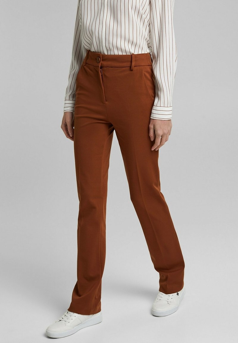 Hnědé kalhoty na míru vyrobené z hladkého materiálu, s zipovým zapínáním, knoflíkovým uzávěrem a předními kapsami, kombinované s bílými teniskami.