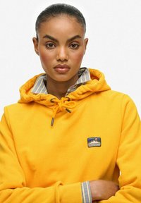 Geel hoodie met trekkoorden en een logopatch; heeft een grijs en wit gestreepte binnenkraag; gladde textuur en casual ontwerp.
