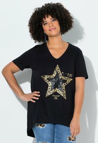 Femme aux cheveux bouclés portant un T-shirt noir à encolure en V avec une grande étoile dorée et noire ainsi que du texte, associée à un jean bleu déchiré.