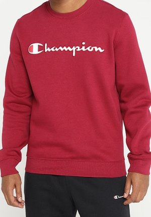 Personne portant un sweat-shirt rouge Champion avec un logo blanc et un pantalon noir avec un petit logo Champion près de la poche.