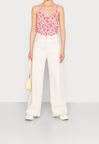 Camiseta de tirantes finos en rosa floral, combinada con unos vaqueros anchos blancos. Sosteniendo un pequeño bolso amarillo brillante. Zapatillas blancas.