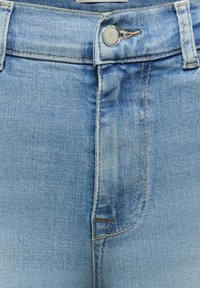 Gros plan sur la ceinture d’un jean en denim bleu clair avec un bouton argenté et des coutures visibles autour de la braguette et des passants de ceinture.
