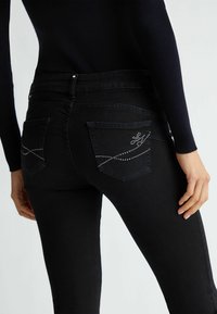 Jeans en denim noir avec une coupe skinny, poches arrière avec un motif décoratif en strass, et une taille mi-haute. Texture lisse.