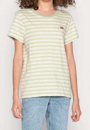 Femme portant un t-shirt Levi's à manches courtes rayé vert clair et blanc, et un jean bleu clair, debout devant un fond uni.