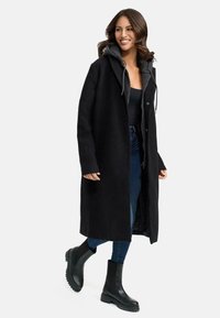 Manteau long noir avec une capuche, doté d'une fermeture éclair à l'avant et de poches latérales, porté sur un haut noir et un jean bleu, accompagné de bottines noires.
