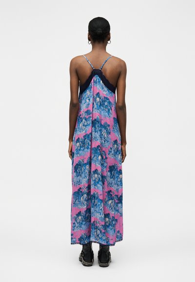 Γυναίκα που φαίνεται από πίσω φοράει μακρύ ροζ και μπλε φόρεμα με λεπτές τιράντες και σκούρο δαντελένιο τελείωμα, σε συνδυασμό με μαύρα χοντρά αθλητικά παπούτσια.