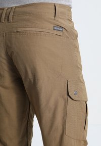 Tan cargo pants made of woven fabric. Features a side pocket with a snap button and a small brand tag on the back pocket. Textured surface.