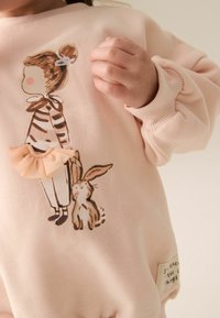 Ljusrosa sweatshirt med ribbad rund halsringning, som har en illustration av en flicka och en kanin, accentuerad med en naken tulle-bow.