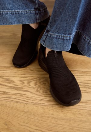 Fødder iført sorte slip-on strikkede sko og blå jeans, stående på et trægulv.